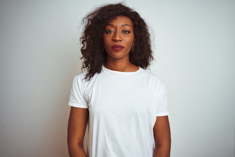 a woman of African decent stands in front of a white wall. She wears a white tee shirt, and has a straight facial expression.