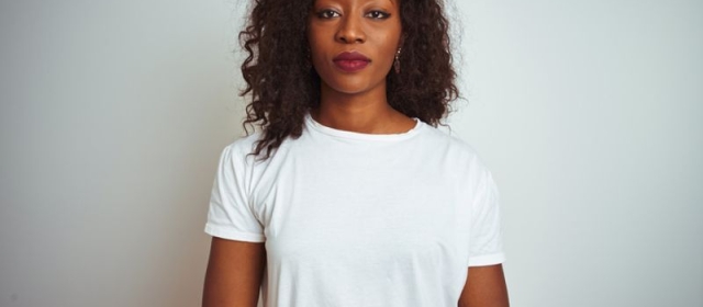 a woman of African decent stands in front of a white wall. She wears a white tee shirt, and has a straight facial expression.