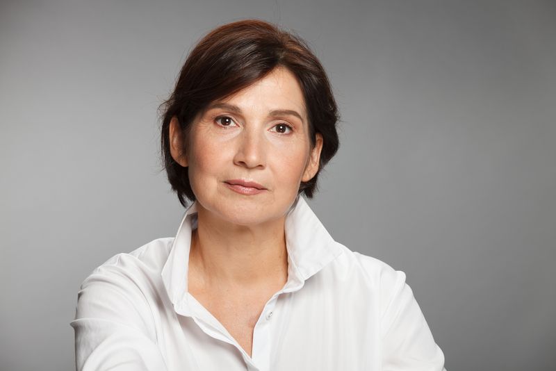 A middle aged woman with short brown hair is pictured. She wears a white collared shirt and is producing a small smile.
