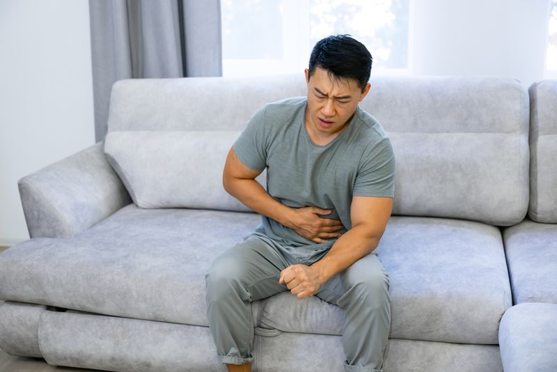A man sits on a gray couch. He wears a blue shirt with gray pants. His face shows distress, as his hand holds his stomach.