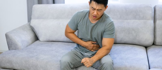 A man sits on a gray couch. He wears a blue shirt with gray pants. His face shows distress, as his hand holds his stomach.