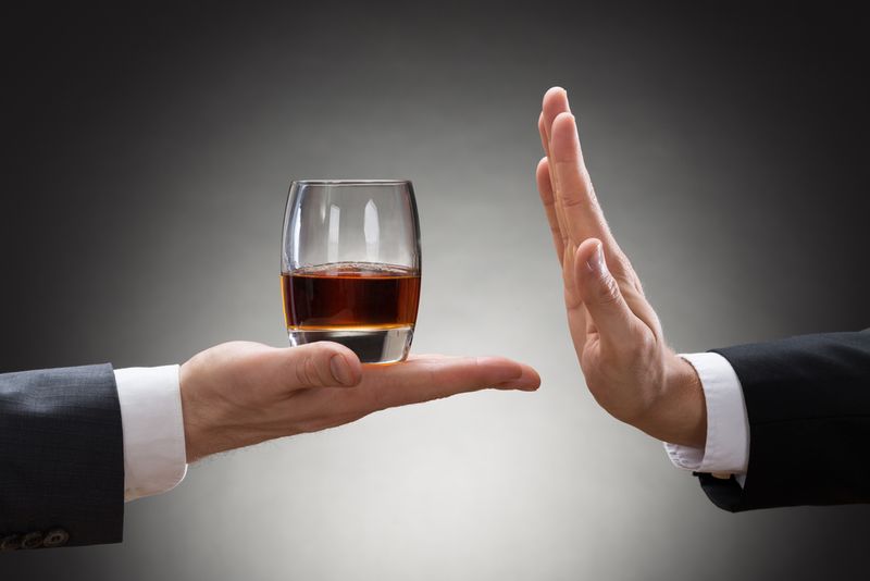 Two hands are pictured. One hand is holding out a glass of whiskey to the other. The other hand is holding itself out in the flat position, to signal that they do not want what is being offered. 