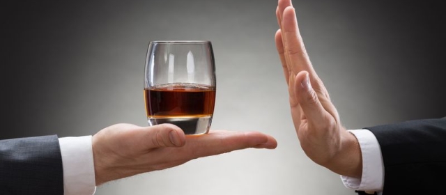 Two hands are pictured. One hand is holding out a glass of whiskey to the other. The other hand is holding itself out in the flat position, to signal that they do not want what is being offered. 