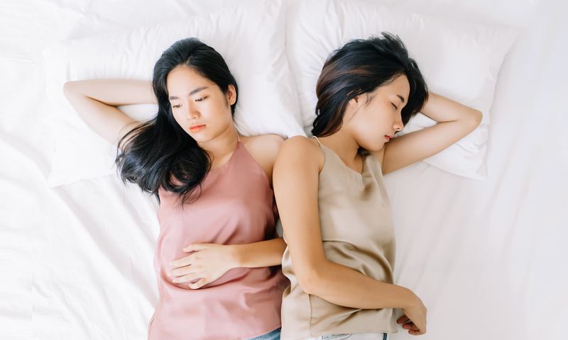 Two women of Asian descent lay next to each other on white sheets. Both wear tank tops. One wears pink, the other wears tan. Both women are looking in opposite directions. 