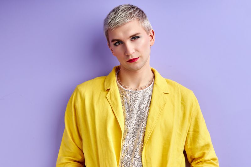 A man stands in front of a purple wall. He wears a sparkling white shirt with a yellow blazer. He wears eyeliner and lipstick.