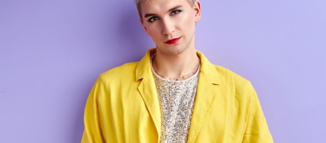 A man stands in front of a purple wall. He wears a sparkling white shirt with a yellow blazer. He wears eyeliner and lipstick.