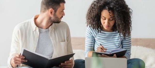 A couple is pictured. The man has short hair, and is wearing a jacket. He sits next to a woman and he is reading a book. The woman is not looking at the man, she is writing in her book instead. the woman has curly hair and wears a blue striped shirt.