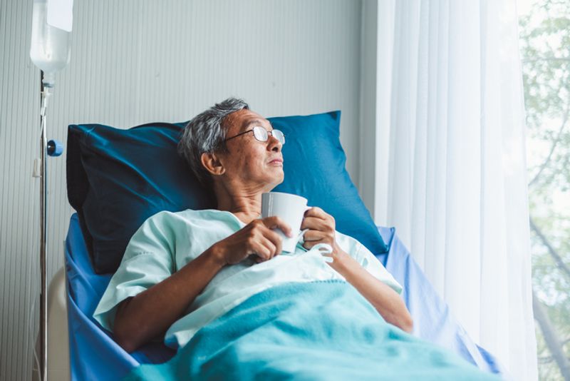 An old man lays in a hospital bed. He is wearing a blue hospital gown, covered in a blue blanket, and is using a blue pillow. He holds a coffee mug, and looks out the window.