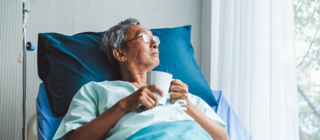 An old man lays in a hospital bed. He is wearing a blue hospital gown, covered in a blue blanket, and is using a blue pillow. He holds a coffee mug, and looks out the window.