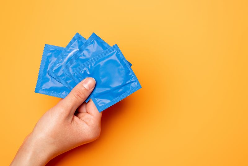 A yellow background reveals a hand holding four blue condoms in wrappers.