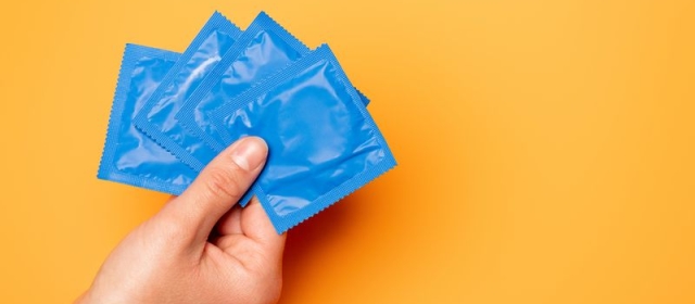 A yellow background reveals a hand holding four blue condoms in wrappers.