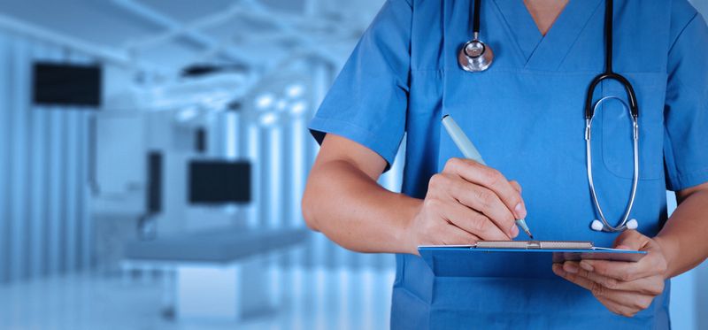 A medical professional is seen from the neck to the waist on the left side of their body. They are wearing blue scrubs with a stethoscope on their neck. Their elbow is extended at a right angle as they use a pen to write on a clipboard.