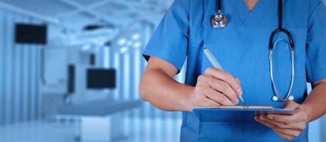 A medical professional is seen from the neck to the waist on the left side of their body. They are wearing blue scrubs with a stethoscope on their neck. Their elbow is extended at a right angle as they use a pen to write on a clipboard.