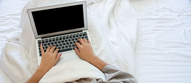 A person, only visible from the chest down, is viewed in their bed. They are covered in a white blanket, sitting on white sheets. They are using a laptop.