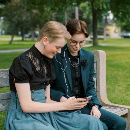Two young LGBTQ+ individuals sitting outside on a bench sharing earbuds and looking at a phone together