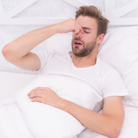 Man in white T-shirt lying in a white bed with fingers pinching his nose in distress