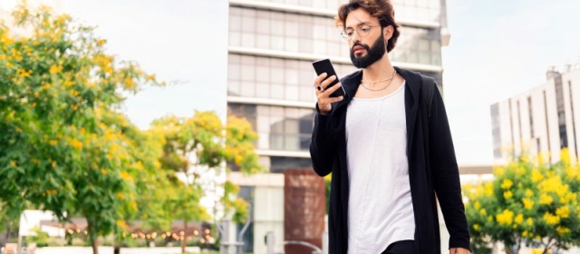 Man with beard walking outside typing on smartphone, wearing white shirt and long black sweater