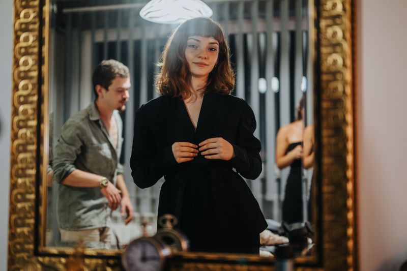 Brown haired woman with bangs gazing at herself in gold-framed mirror wearing lack suit jacket