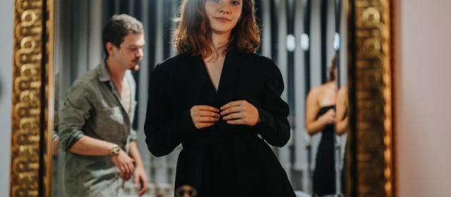 Brown haired woman with bangs gazing at herself in gold-framed mirror wearing lack suit jacket