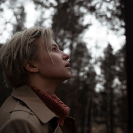 Blond, short-haired person with orange turtleneck sweater and tan trench coat staring at sky intensely in a forested location