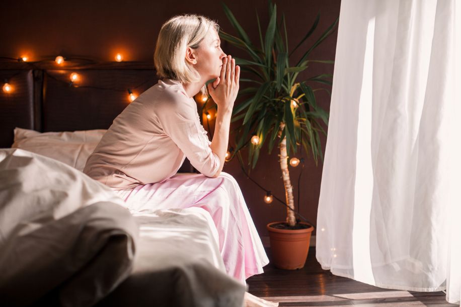 Postmenopausal woman sitting on the edge of her bed in pink pajamas, with hands in a prayer position to her lips. Contemplating life