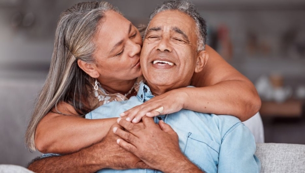 older woman hugging older man from behind and cheek kiss, older man smiling