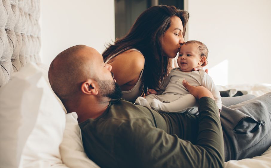 dad laying down holding baby, mom leans over and kisses baby on forehead